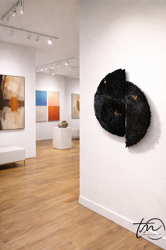 Art gallery with a black abstract sculpture on a white wall and colorful paintings in the background. Original textured abstract wall art by Artist Tam M Olson
