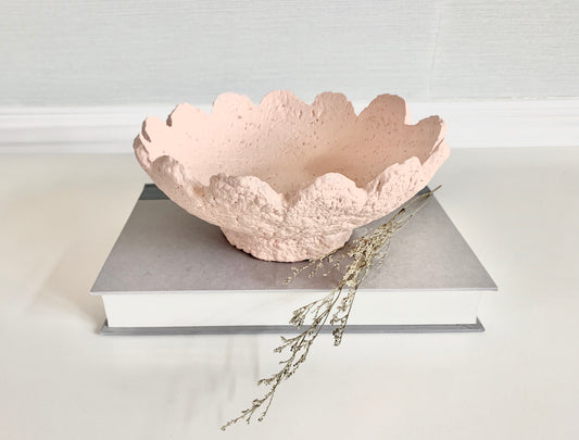 A soft pink paper mache bowl with scalloped wavy details, displayed on a book with a few dried flowers, set against a neutral background. Handmade by artist Tam M Olson.