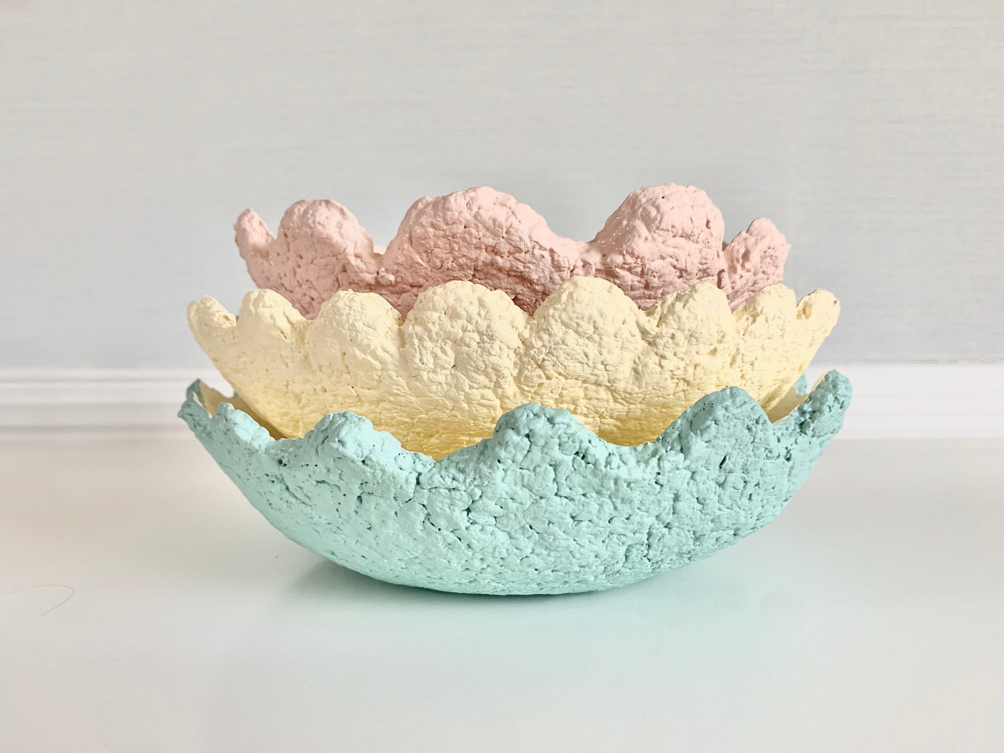 Three pastel colored wavy scalloped paper mache bowls nested inside of each other. The bottom bowl is teal with wavy edging. The middle bowl is yellow with scallop edging. The top bowl is pink with wavy scallop edging. Handcrafted by artist Tam M Olson.