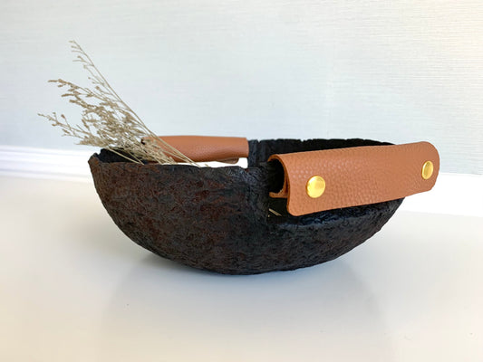 A rustic black paper mache bowl with leather handles, displaying a natural aged appearance and leather snap handles. Hand crafted by Artist Tam M Olson.