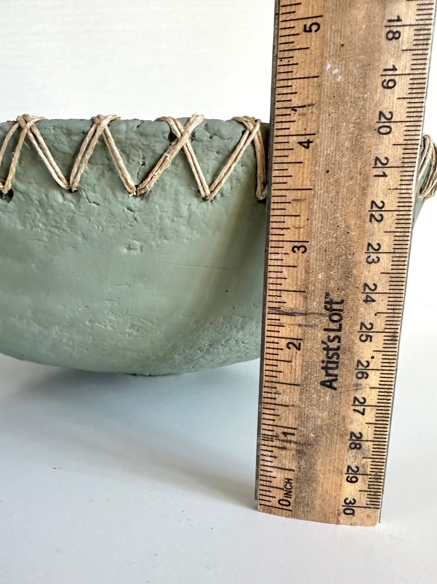 Decorative soft green bowl with woven edge next to a wooden ruler on a white background. Ruler displays a height of about 4.25 inches. Bowl is handmde by Artist Tam M Olson