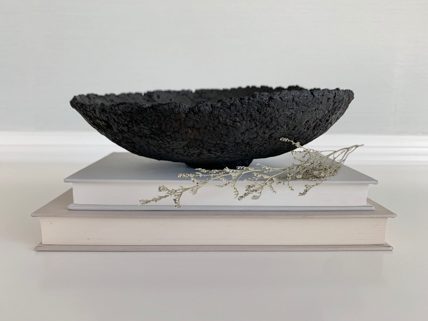 A rustic black decorative bowl made with paper mache, displayed on a white shelf with books, against a neutral background. Designed by Artist Tam M Olson.