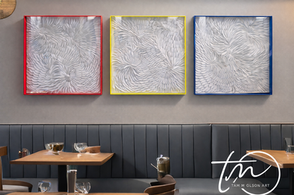 Three white abstract dimensional artworks in transparent hot pink, neon yellow, and blue frames on a gray wall above a restaurant booth. Original Art by artist Tam M Olson.