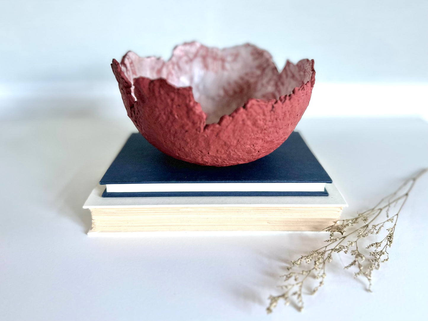 A textured maroon eggshell decorative bowl with a rough torn edge, placed a top books. Handcrafted by artist Tam M Olson.