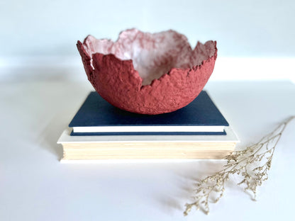 A textured maroon eggshell decorative bowl with a rough torn edge, placed a top books. Handcrafted by artist Tam M Olson.