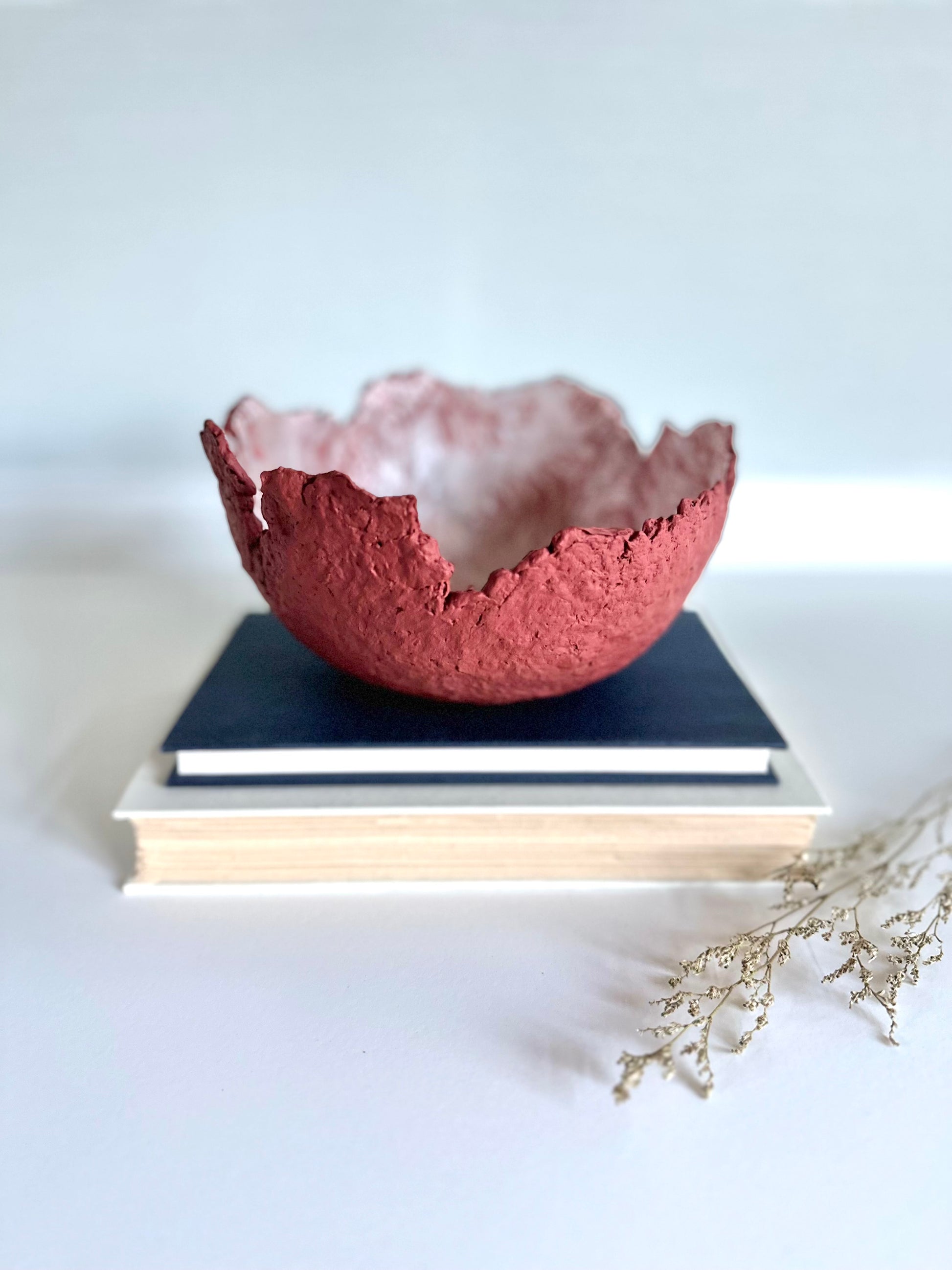 A textured maroon eggshell decorative bowl with a rough torn edge, placed a top books. Handcrafted by artist Tam M Olson.