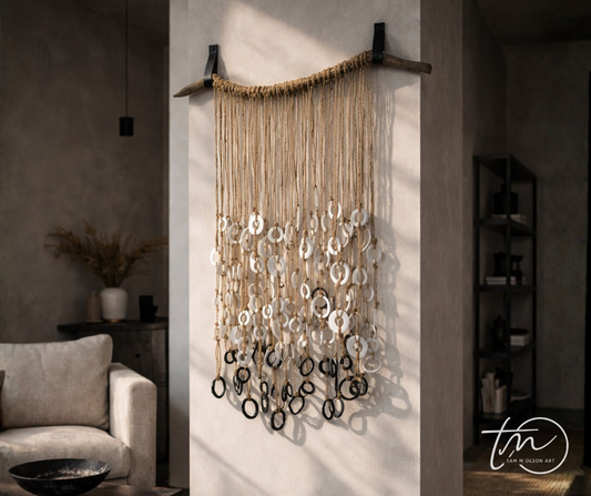 A wall hanging decor made of recycled driftwood with leather straps, jute string, and clay rings, sealed with paint and coffee grounds. Abstract artwork hanging on narrow wall in organic modern living space. Original artwork by artist Tam M Olson.