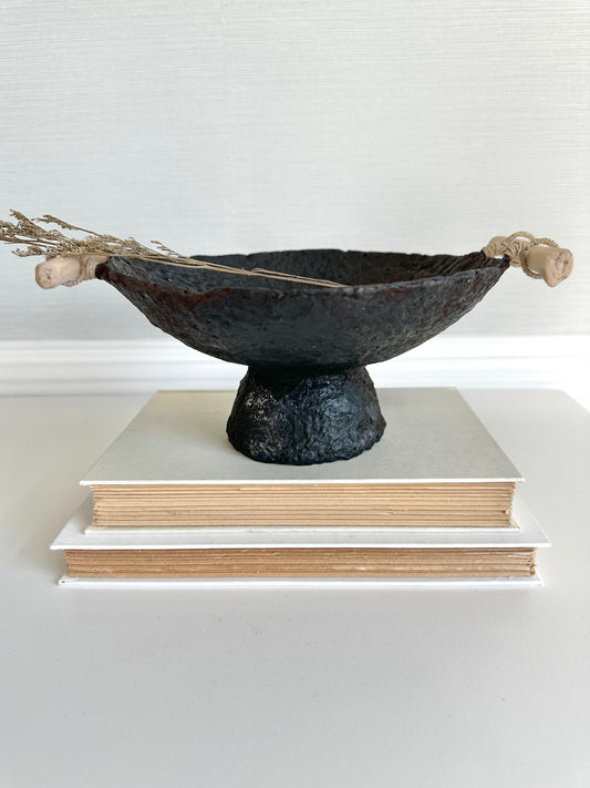 A black pedestal bowl made of paper mache, featuring a rustic and textured design, displayed on a stack of books. Handcrafted by Artist Tam M Olson.