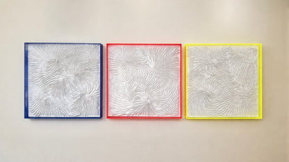 Three abstract artworks with textured patterns in blue, red, and yellow frames on a beige wall. Art by artist Tam M Olson