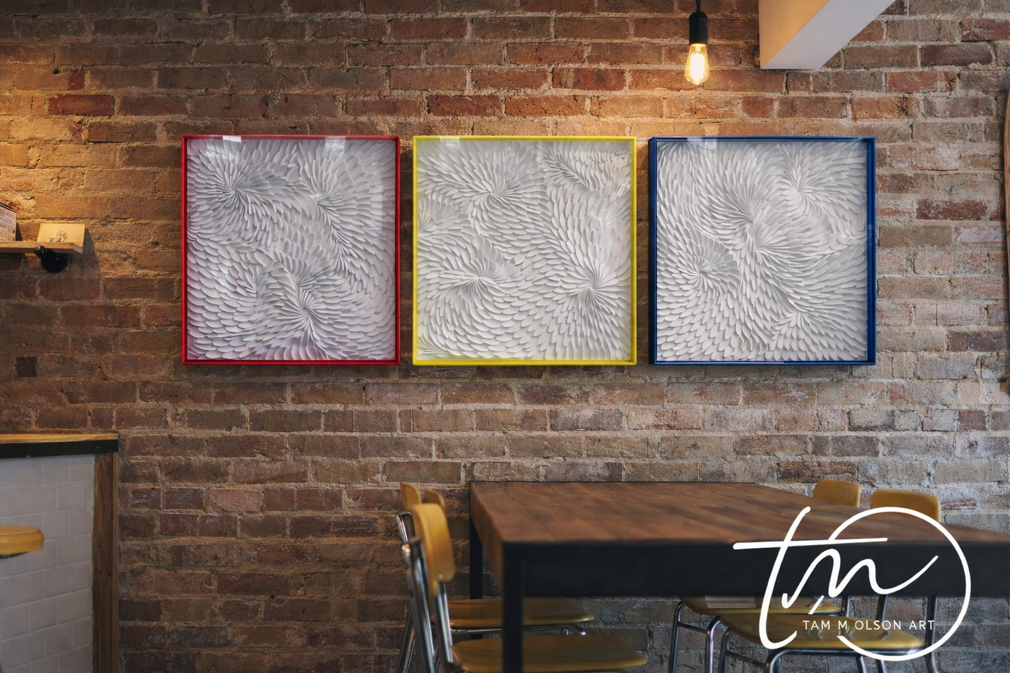 Three white textured wall art pieces in transparent hot pink, neon yellow, and blue frames on a brick wall with a dining table and chairs below. Original art by Artist Tam M Olson