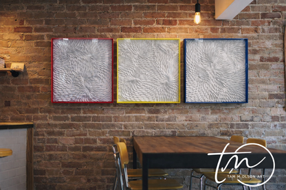 Three white textured wall art pieces in transparent hot pink, neon yellow, and blue frames on a brick wall with a dining table and chairs below. Original art by Artist Tam M Olson