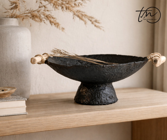 A black pedestal bowl with a patina brown finish and driftwood handles on a table with other minimal decor items. Handcrafted by Artist Tam M Olson.