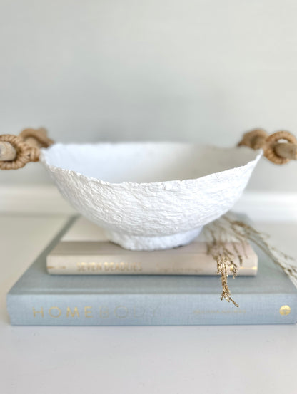 A handmade white paper mache bowl with driftwood and jute handles as a decorative item, placed on a stack of books. Designed by Artist Tam M Olson.