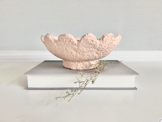 A soft pink paper mache bowl with scalloped wavy details, displayed on a book with a few dried flowers, set against a neutral background. Handmade by artist Tam M Olson.