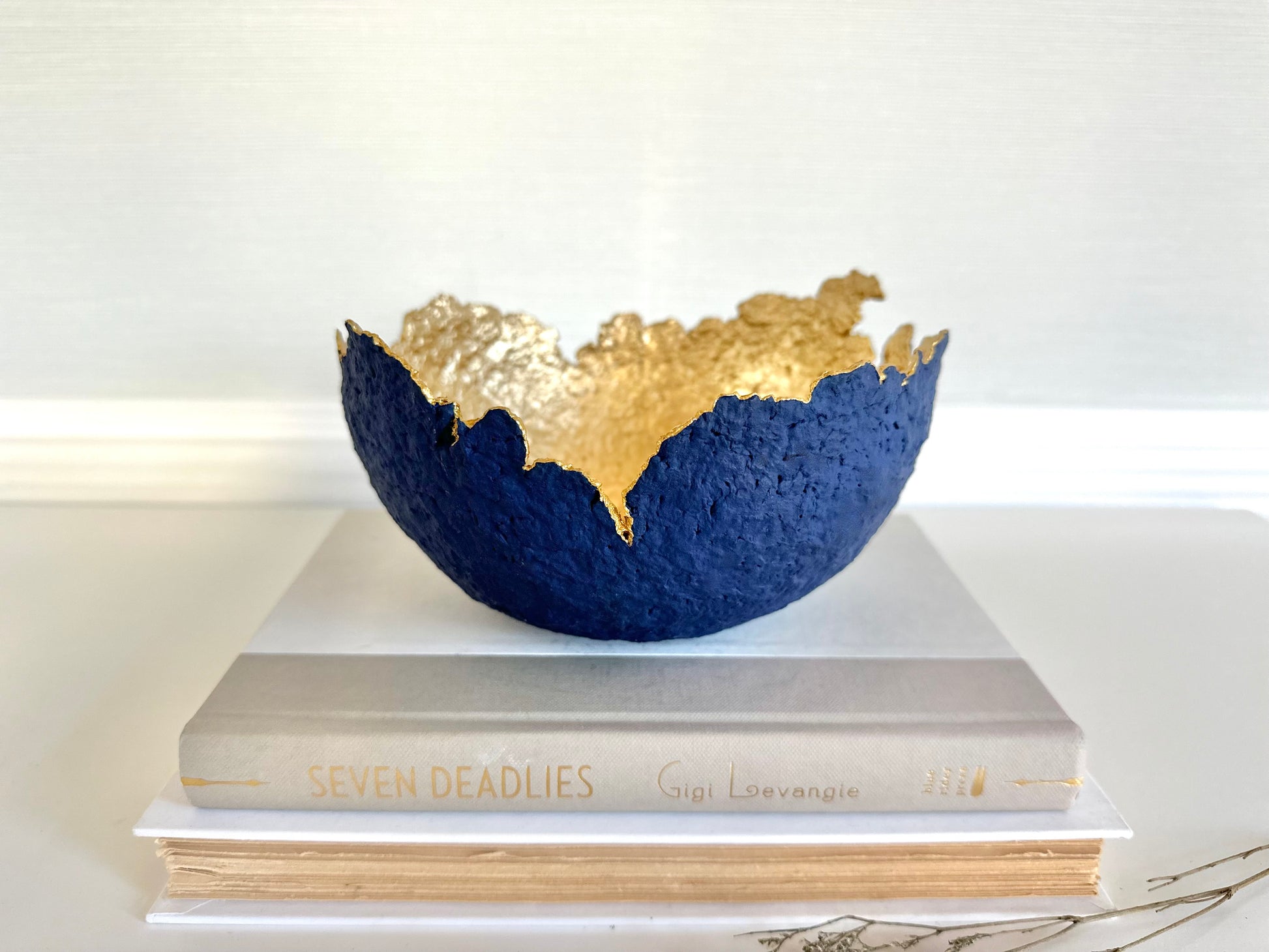 A navy blue paper mache bowl with a gold interior, designed to resemble an eggshell, placed on a white surface with a book and decorative dry branches nearby. Original sculptural bowl by Artist Tam M Olson