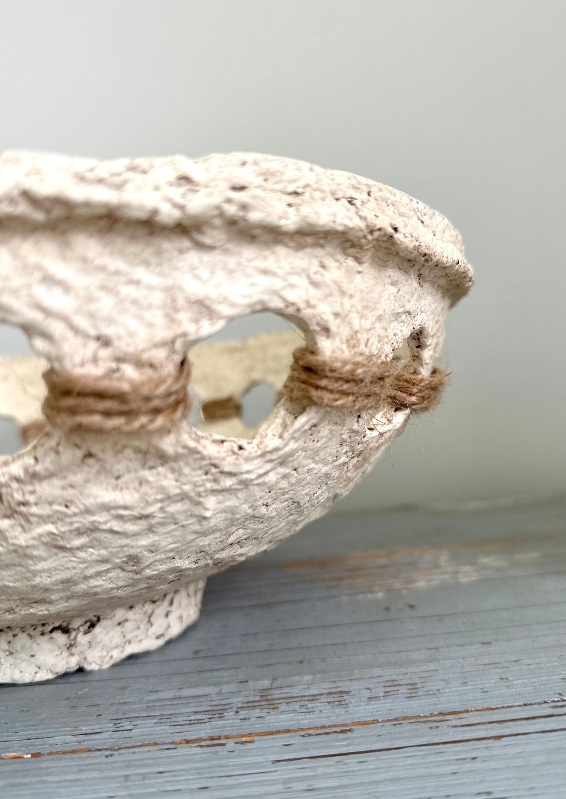 Close up of a beige, primitive handmade paper mache bowl with jute string accents on the side, placed on a flat surface. Designed by Artist Tam M Olson.