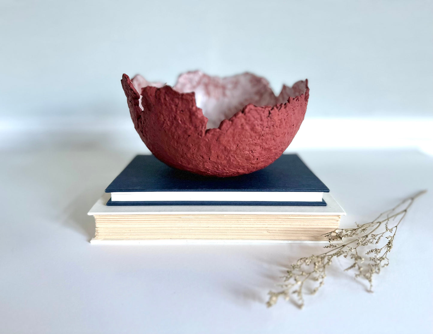 A textured maroon eggshell decorative bowl with a rough torn edge, placed a top books. Handcrafted by artist Tam M Olson.