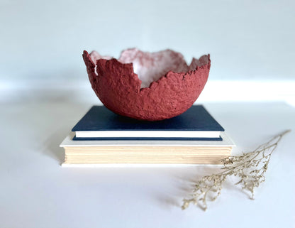 A textured maroon eggshell decorative bowl with a rough torn edge, placed a top books. Handcrafted by artist Tam M Olson.