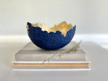 A navy blue paper mache bowl with a gold interior, designed to resemble an eggshell, placed on a white surface with a book and decorative dry branches nearby. Original sculptural bowl by Artist Tam M Olson