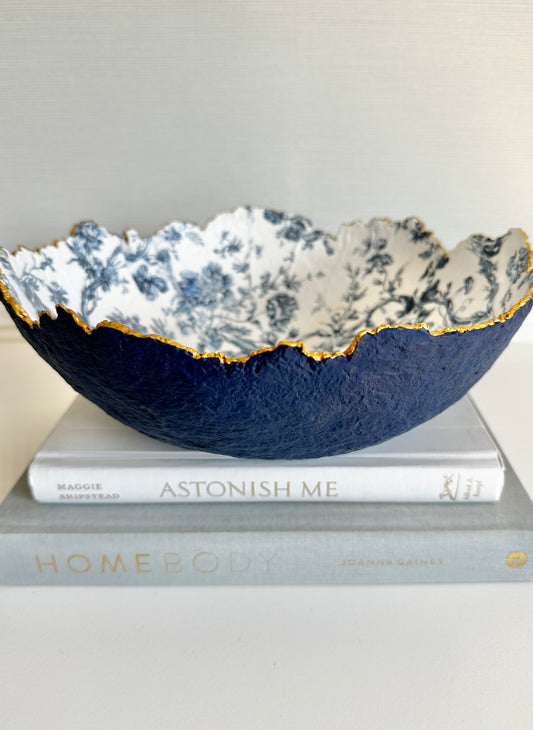A navy blue textured bowl with irregular gold edging and a floral toile pattern on the inside, placed on two books. Handcrafted paper mache bowl by artist Tam M Olson.