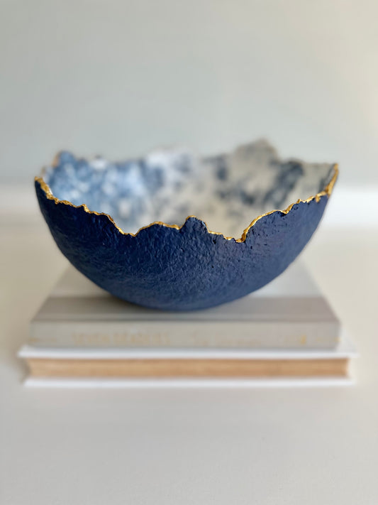 A navy blue textured bowl with irregular gold edging and a floral toile pattern on the inside, placed on two books. Handcrafted paper mache bowl by artist Tam M Olson.