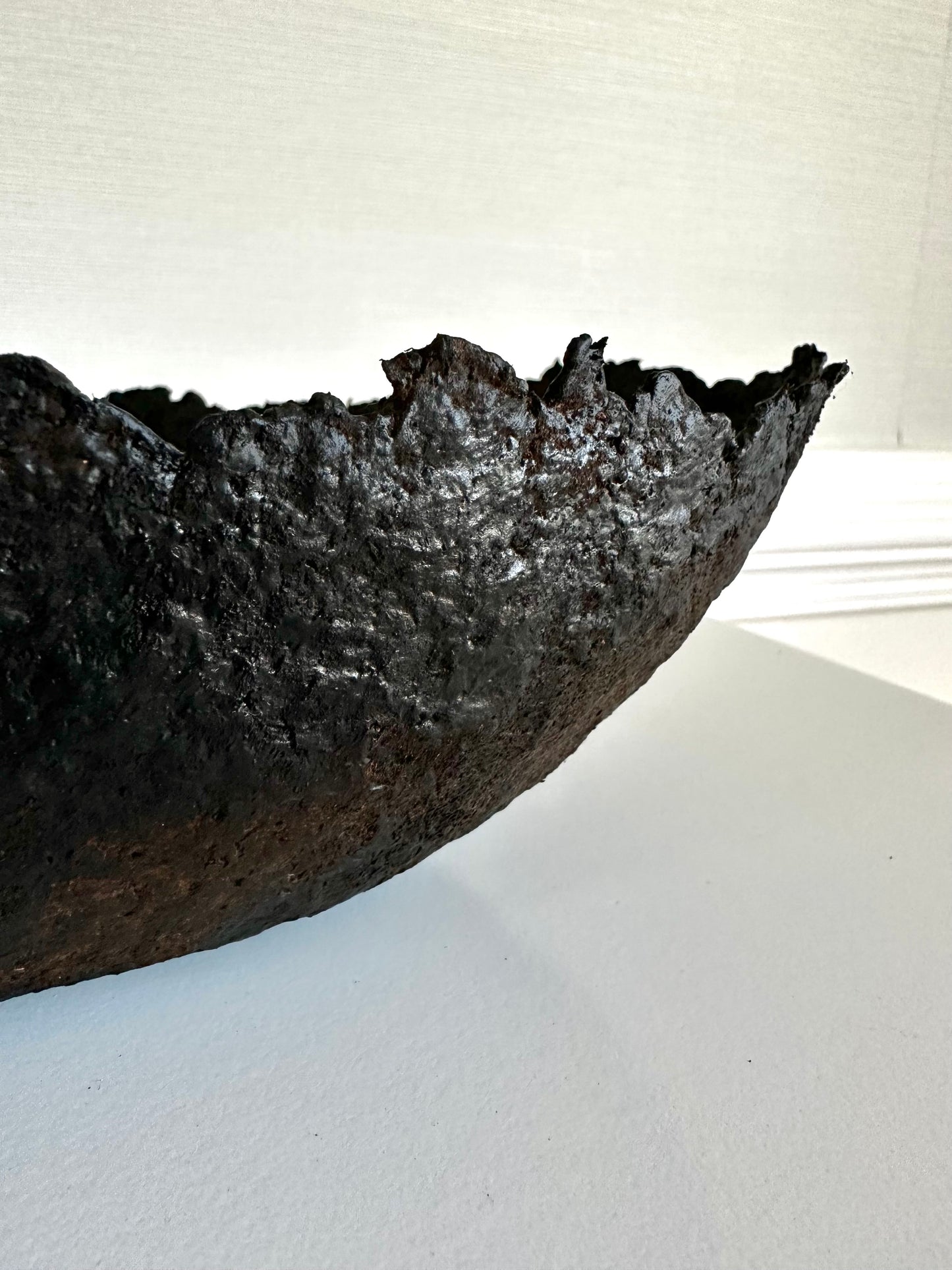 Close up of oblong black textured decorative bowl with irregular edges on white surface. Handmade Paper Mache design by Artist Tam M Olson