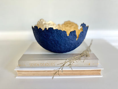A navy blue paper mache bowl with a gold interior, designed to resemble an eggshell, placed on a white surface with a book and decorative dry branches nearby. Original sculptural bowl by Artist Tam M Olson