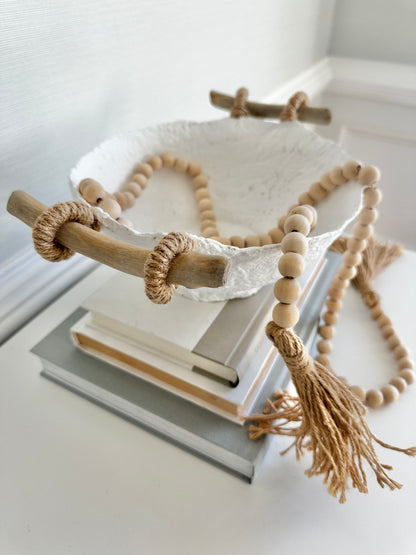 A handmade white paper mache bowl with driftwood and jute handles as a decorative item, filled with wooden strung beads, placed on a stack of books. Designed by Artist Tam M Olson.