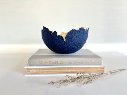 A navy blue paper mache bowl with a gold interior, designed to resemble an eggshell, placed on a white surface with a book and decorative dry branches nearby. Original sculptural bowl by Artist Tam M Olson