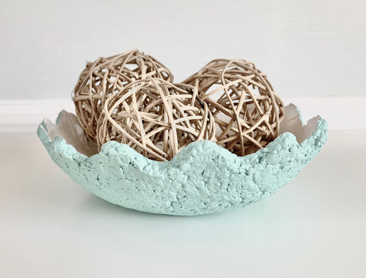 A pastel teal paper mache decorative bowl with wavy edges and a white interior, containing straw ball decorative objects. Handmade by artist Tam M Olson.