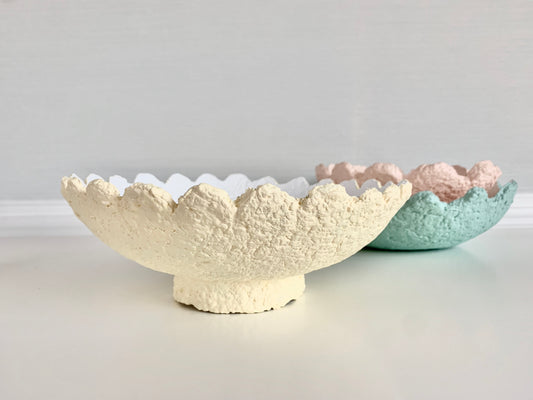 A light yellow scalloped decorative bowl made from paper mache, with wavy edges and a slightly uneven texture, displayed alongside two similar pastel bowls. Original handmade work by artist Tam M Olson