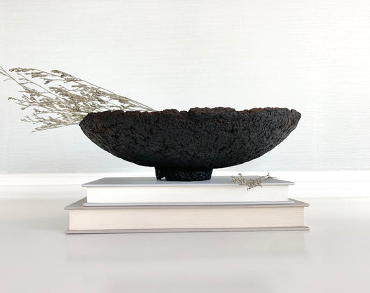 A rustic black decorative bowl made with paper mache, displayed on a white shelf with books, against a neutral background. Designed by Artist Tam M Olson.