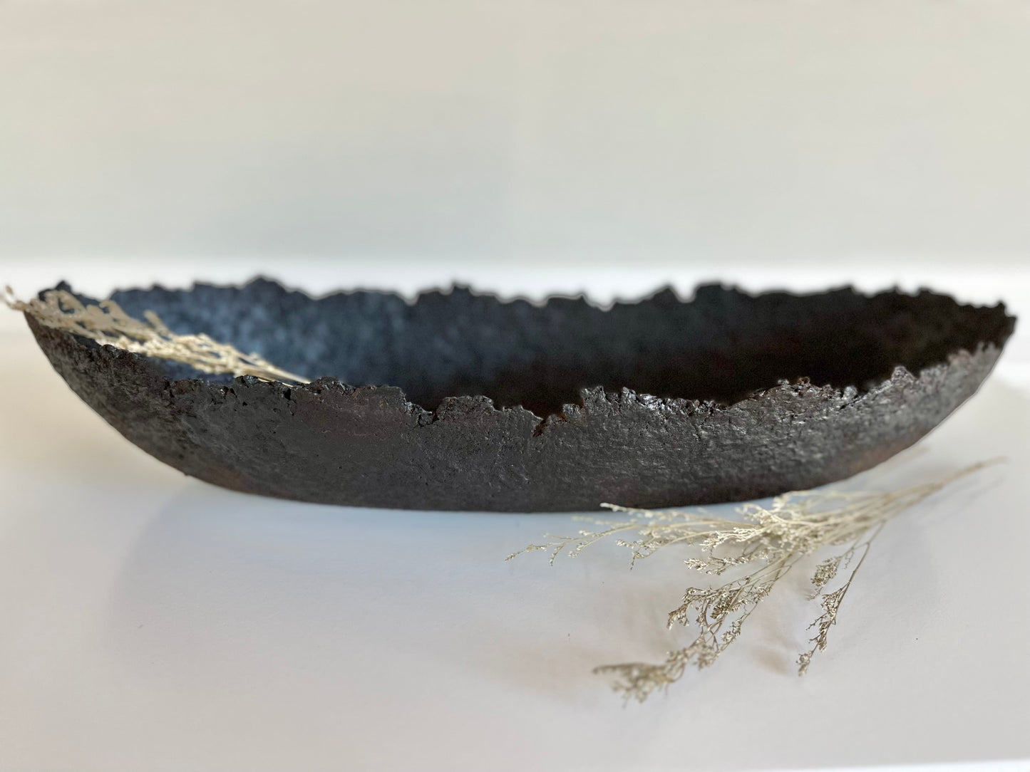 Oblong black textured decorative bowl with irregular edges on white surface. Handmade Paper Mache design by Artist Tam M Olson