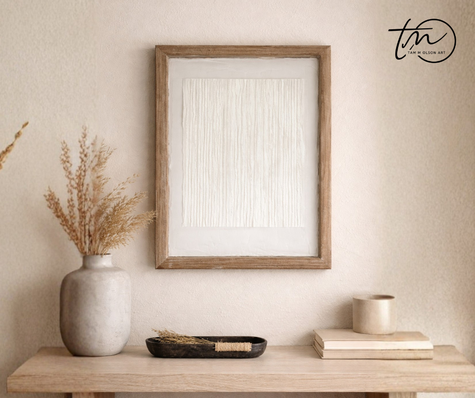 Textured abstract white art in a wooden frame on a shite wall above a table with other decorative objects. Original art by artist Tam M Olson