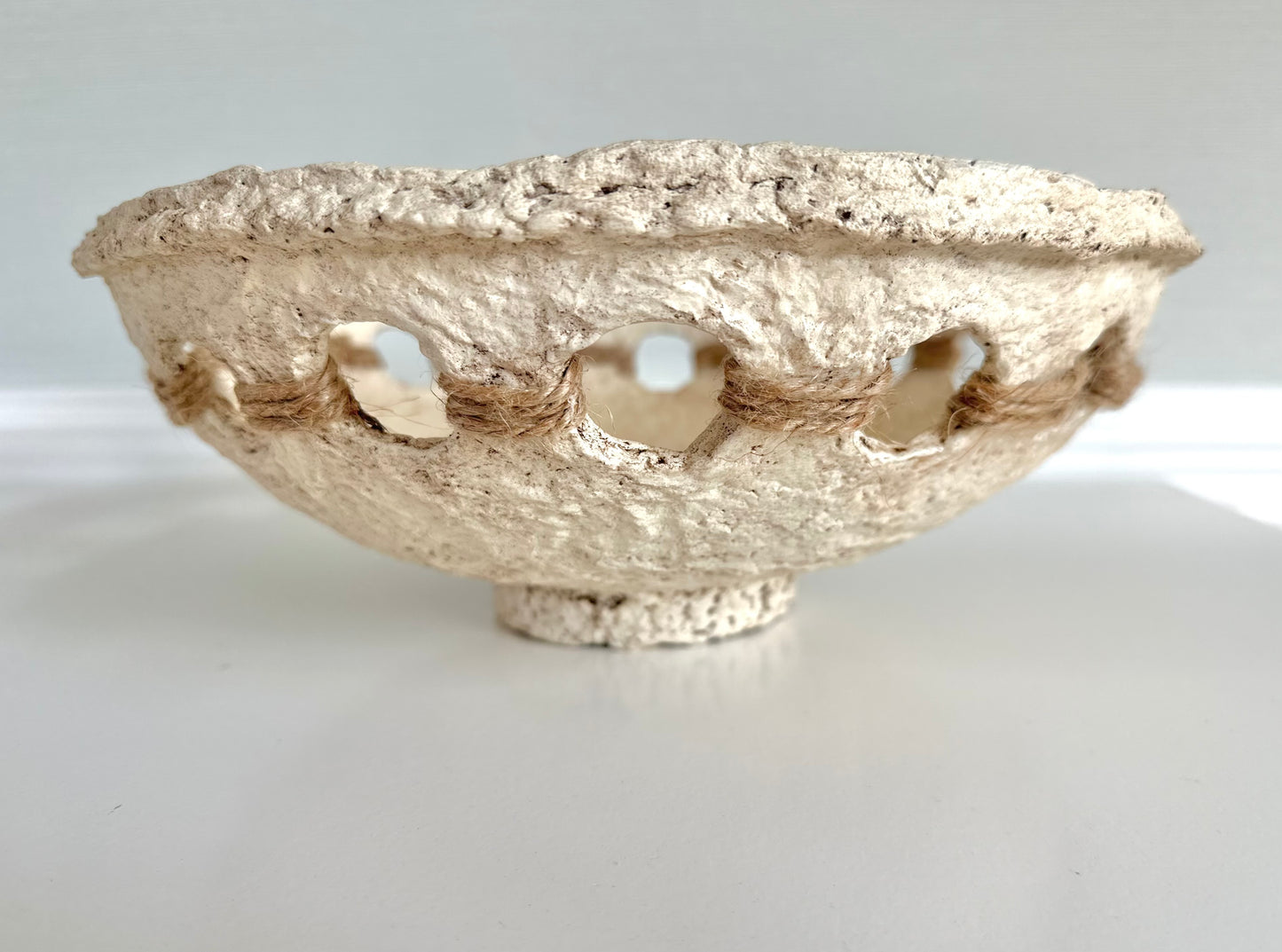 A beige, primitive handmade paper mache bowl with jute string accents on the side, placed on a flat surface. Designed by Artist Tam M Olson.