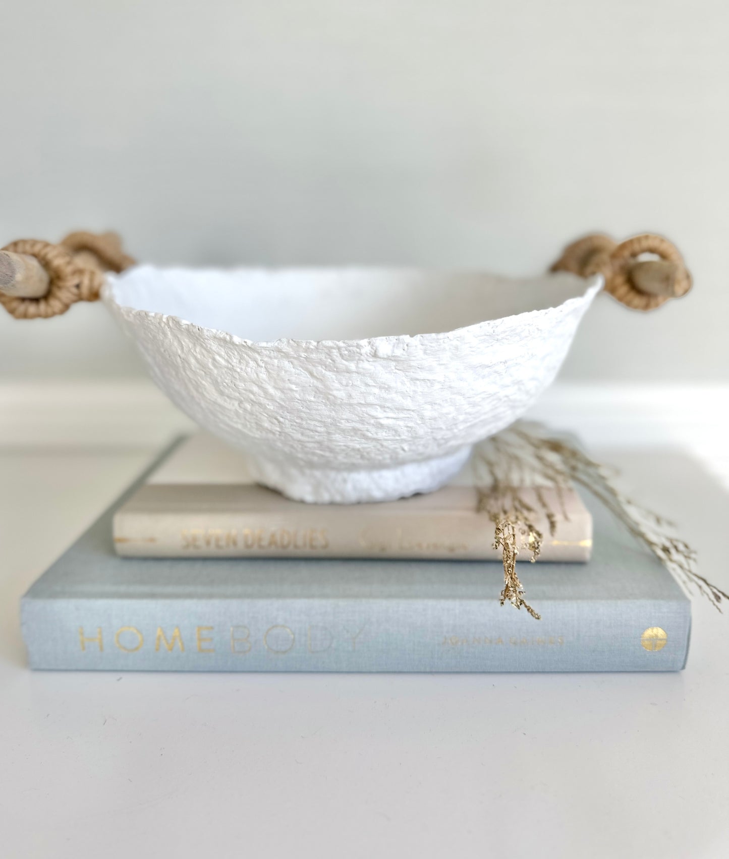 A handmade white paper mache bowl with driftwood and jute handles as a decorative item, placed on a stack of books. Designed by Artist Tam M Olson.