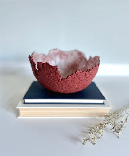 A textured maroon eggshell decorative bowl with a rough torn edge, placed a top books. Handcrafted by artist Tam M Olson.