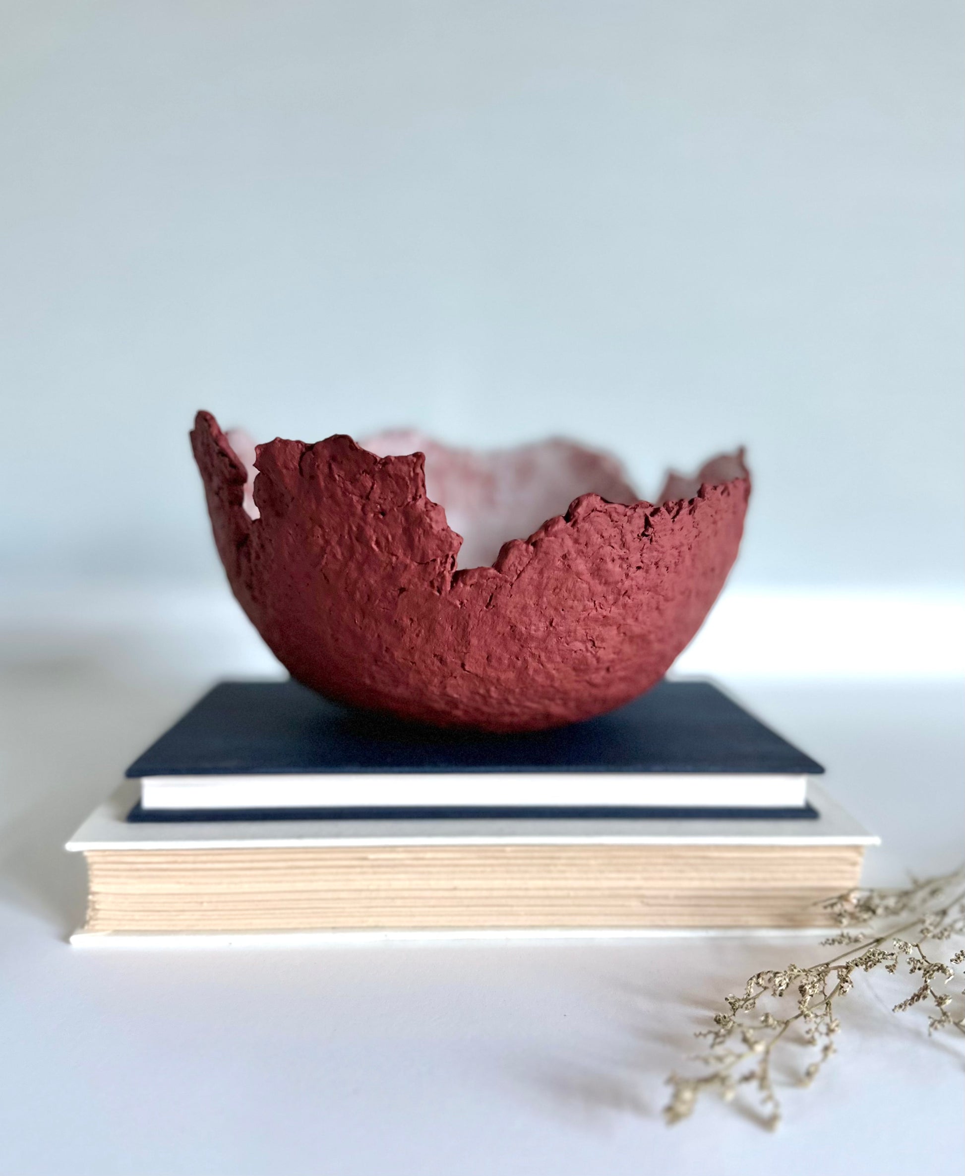 A textured maroon eggshell decorative bowl with a rough torn edge, placed a top books. Handcrafted by artist Tam M Olson.