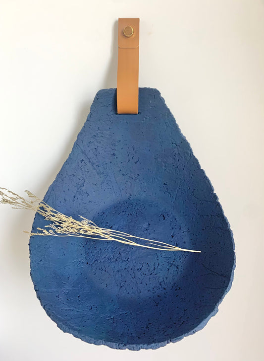 Blue paper mache tear drop sculpture with a leather strap on a white wall, handmade by Artist Tam M Olson