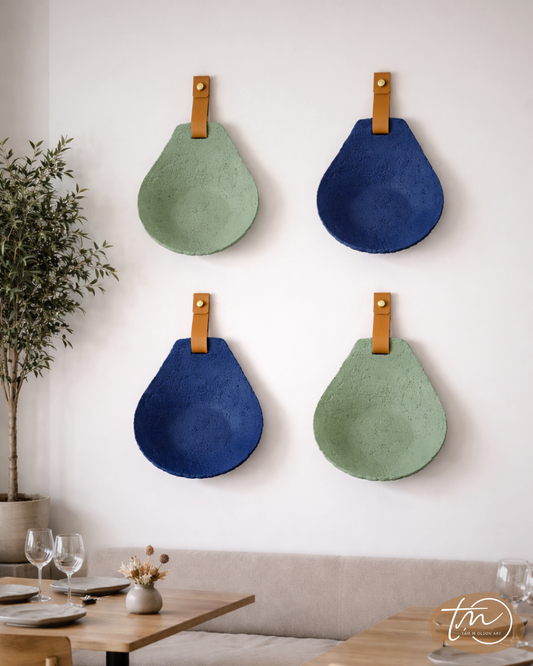 Four wall-mounted paper mache sconces in green and blue on a white wall with a dining area in the background. Handmade by Artist Tam M Olson