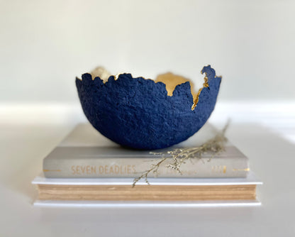 A navy blue paper mache bowl with a gold interior, designed to resemble an eggshell, placed on a white surface with a book and decorative dry branches nearby. Original sculptural bowl by Artist Tam M Olson