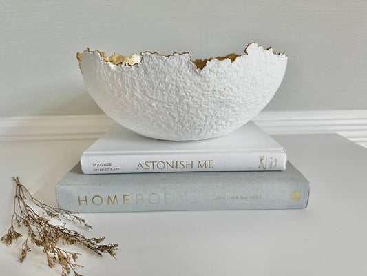 White Decorative textured bowl with  irregular gold edge made with paper mache material placed on stacked books. Original bowl handmade by artist Tam M Olson